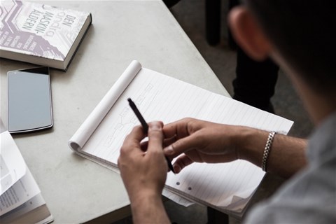 Bild som visar en students händer vid ett bord, med penna, kursbok, block och mobiltelefon
