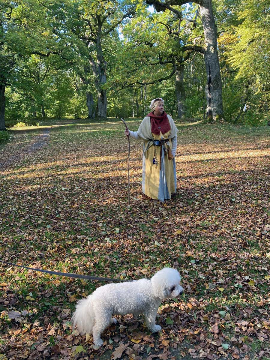 Kvinna i medeltidskläder i en park. En hund står framför henne